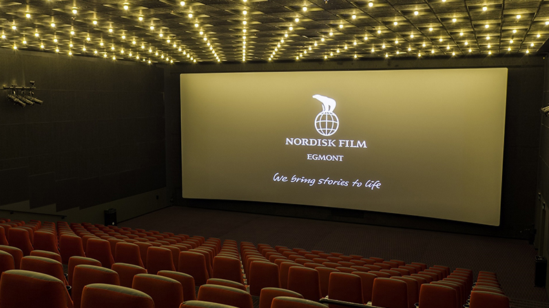 En tom Nordiske Film biografsal med guld belysning. På biograflærred står "We bring stories to life"