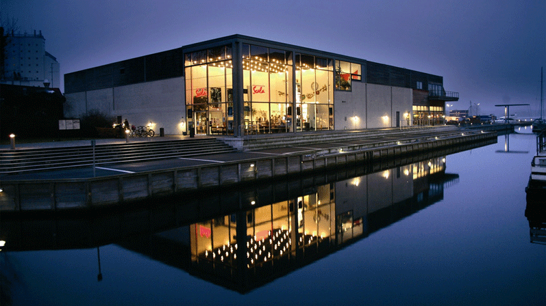 Stor og moderne bygning ved vandet med et stort glasparti i midten og gæster indenfor