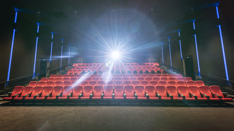 Foran en stor og tom biograf med røde siddepladser, blå væglamper og en meget skarp, lysende projektor