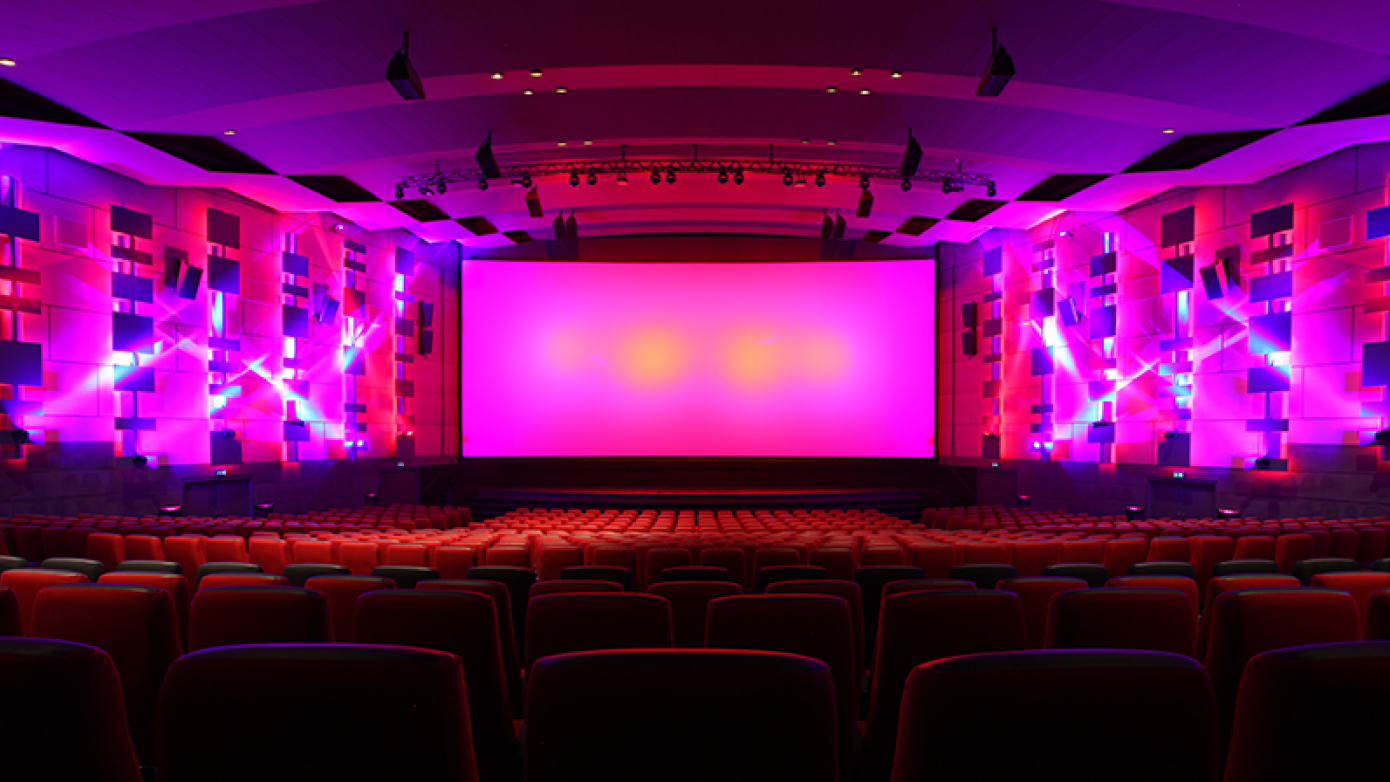 Meget stor biografsal med mange tomme biografsæder. Rummet er belyst med skarpe lyserøde projektorer
